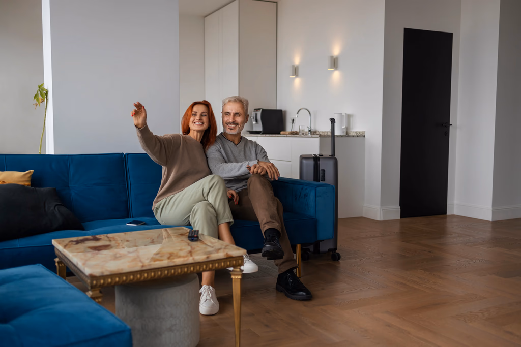 A happy senior couple relaxing on a blue sofa in a spotless, well-lit living room, appearing satisfied with their home environment.
