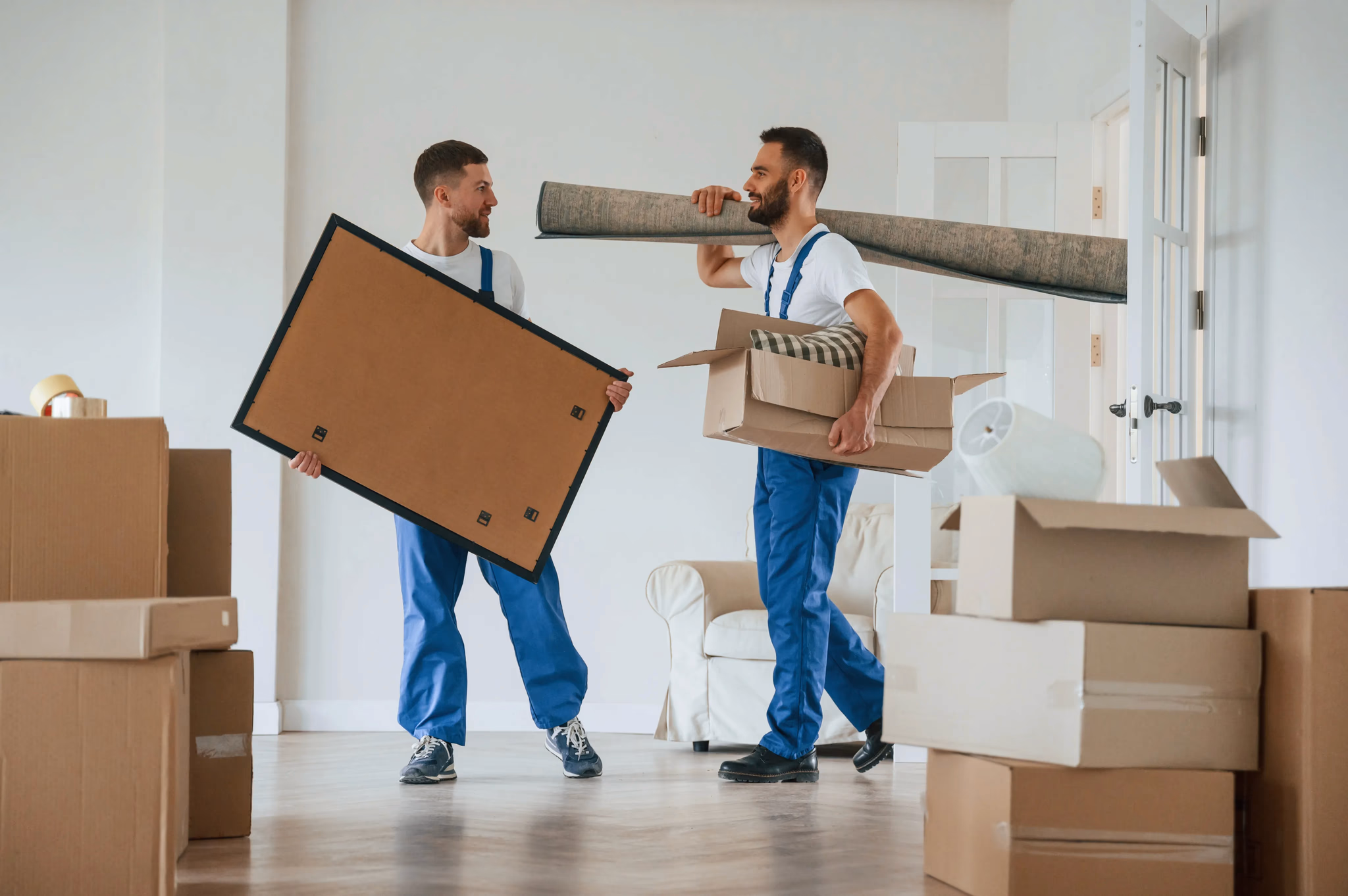 Two professional cleaners carrying a rolled rug and cardboard boxes in a bright room, showcasing move-in and move-out cleaning services for new tenants.